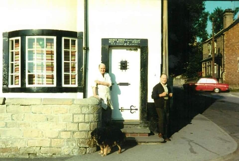 Two men standing on steps in front of a shop on the corner of a street with their dog.